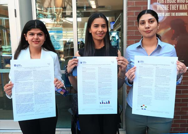 Three JIBC students holding up their research papers with a blue table in front of them.
