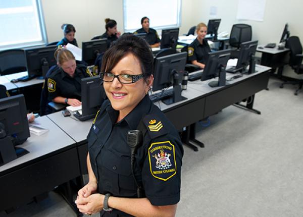 A corrections officer stands in front of a group of co-workers, each working at computers, in a professional setting.
