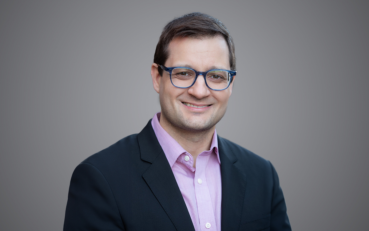 Head and shoulders photo of man in dark jacket and magenta shirt wearing glasses.