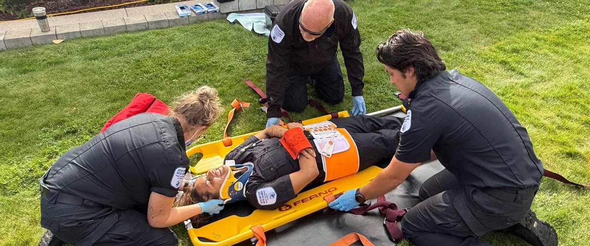 Three paramedic students in black uniforms secure a fourth student onto a stretcher.
