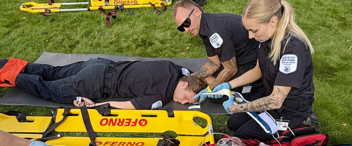 A male paramedic student with hands under the neck of another male paramedic student lying face down on a stretcher on the grass while a female paramedic student works with a bandage.