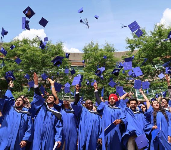 JIBC graduates in blue gowns toss their blue grad caps in the air.