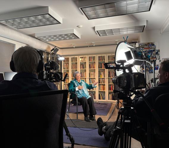 Woman sits in chair gesturing as two men sit behind lights and video cameras pointed at her.n