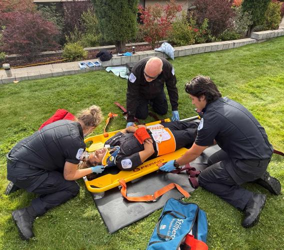 Three paramedic students in black uniforms secure a fourth student onto a stretcher.
