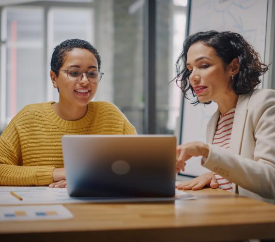 A woman gestures to a laptop screen while another woman looks on.