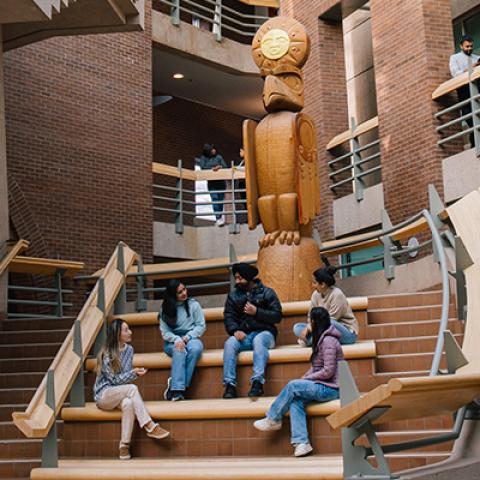 Five students sitting on steps in atrium below Welcome Post at New Westminster campus.