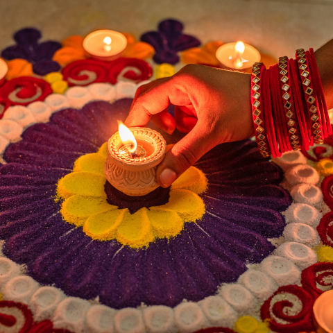 A person places a candle with their right hand onto a vibrant rangoli design crafted with swirling patterns of purple, yellow, red, orange, and white.