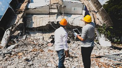 Two relief workers wearing hard hats look at collapsed building.