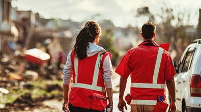 Two relief workers in safety vests walk amidst the wreckage.