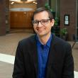 Dark haired man with glasses wearing black jacket and royal blue shirt.