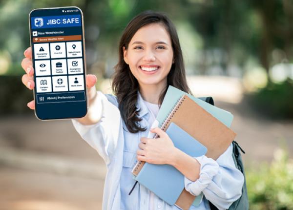 Woman holding notebooks in left hand and holding cell phone with JIBC Safe app on screen with outstretched right hand.