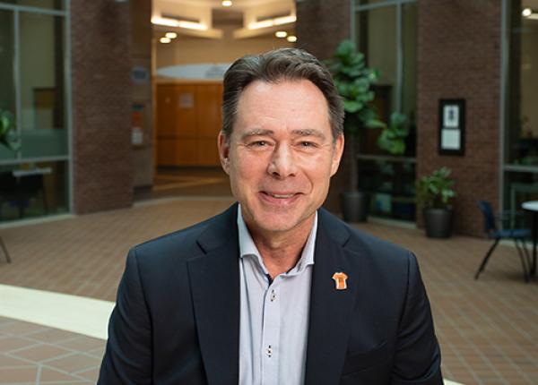 Dark-haired man wearing dark jacket and grey shirt.